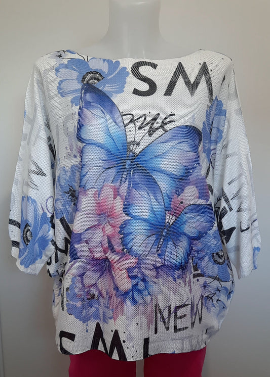 Haut ample en maille blanche à manches 3/4, imprimé papillons bleus et fleurs dans les tons rose et violet, détails de textes en noir façon street art. Style estival, artistique et féminin.