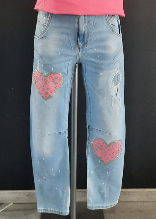 Jean bleu clair pour femme avec cœurs rose léopard pailletés sur les jambes, effet usé et éclaboussures blanches pour un style streetwear fun et décontracté.