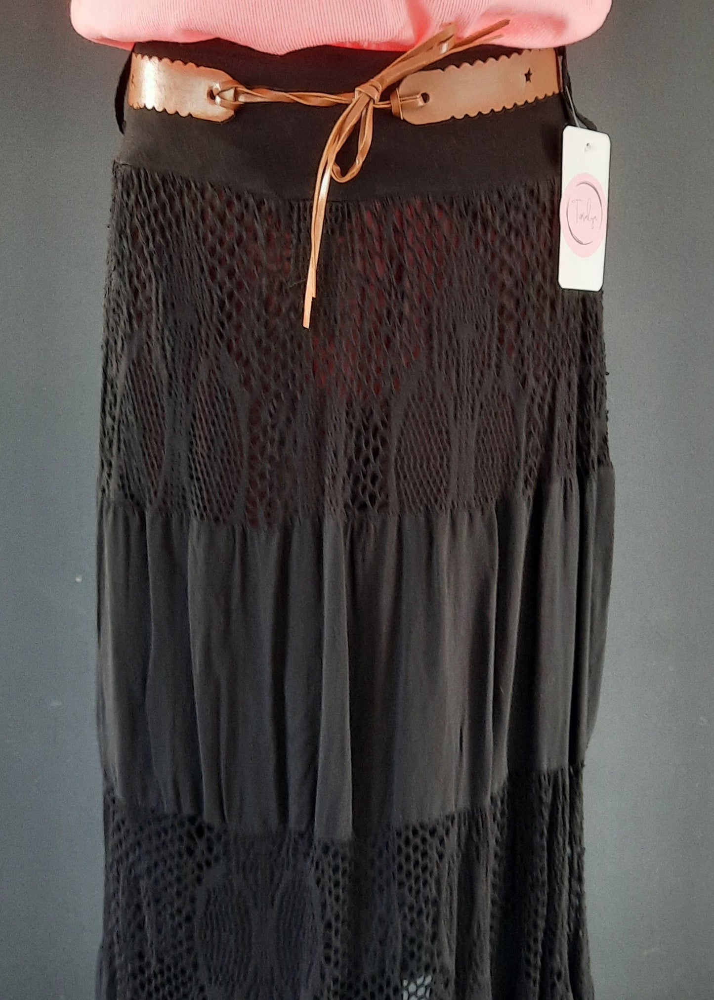 Jupe noire longue à volants, ornée d’empiècements en dentelle ajourée, portée avec une ceinture camel et un haut rose. Style bohème chic, idéale pour la mi-saison.