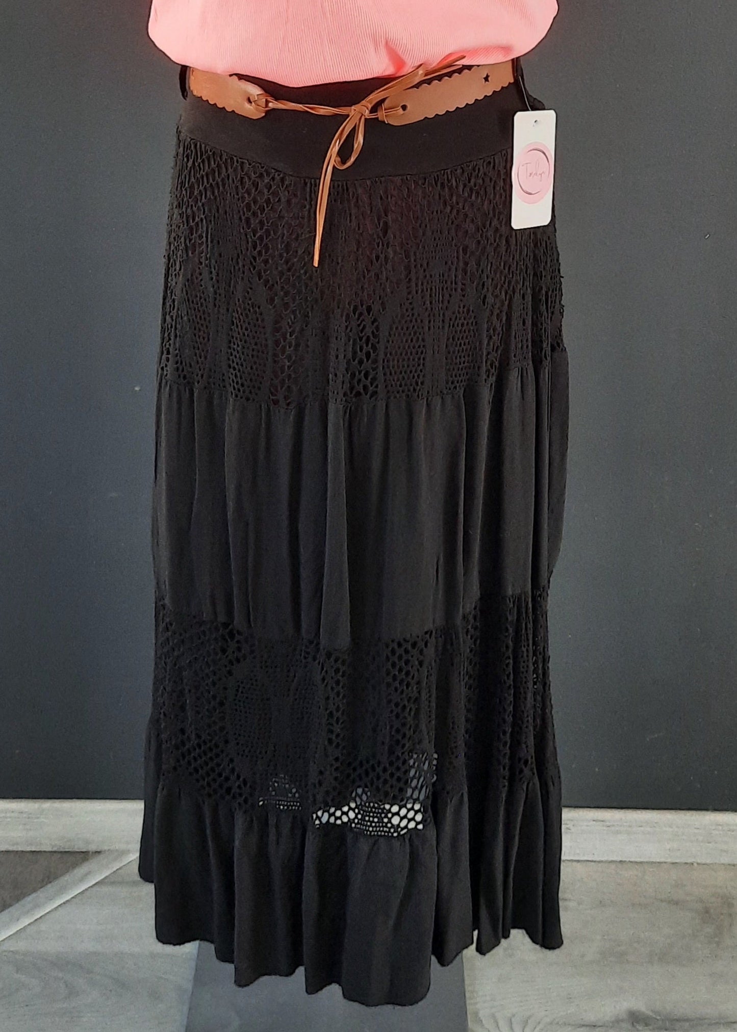 Jupe noire longue à volants, ornée d’empiècements en dentelle ajourée, portée avec une ceinture camel et un haut rose. Style bohème chic, idéale pour la mi-saison.
