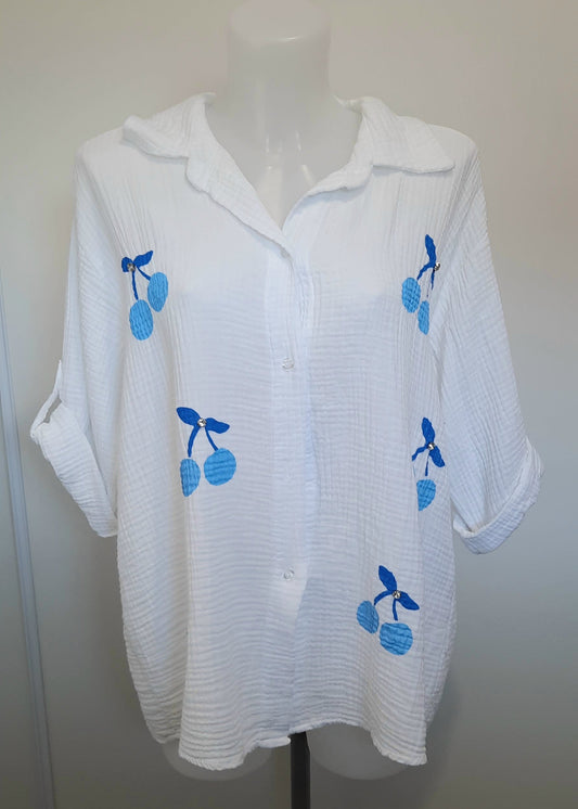Chemise blanche en gaze de coton pour femme, ornée de broderies cerises bleues avec strass – modèle à manches longues retroussables et col classique pour un style estival et ludique.