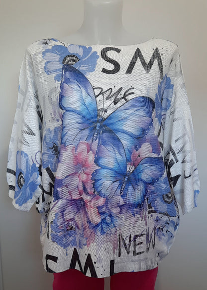 Haut ample en maille blanche à manches 3/4, imprimé papillons bleus et fleurs dans les tons rose et violet, détails de textes en noir façon street art. Style estival, artistique et féminin.