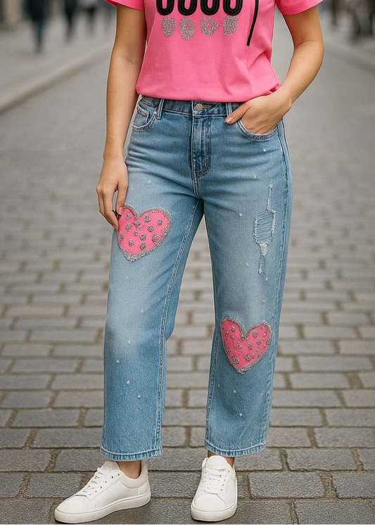 Jean bleu clair pour femme avec cœurs rose léopard pailletés sur les jambes, effet usé et éclaboussures blanches pour un style streetwear fun et décontracté.