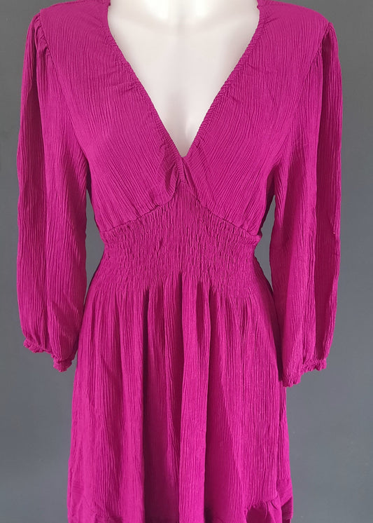 Robe courte fuchsia à manches longues, en tissu gaufré, avec un décolleté cache-cœur et une taille smockée. Coupe fluide et féminine, idéale pour le printemps ou l’été.
