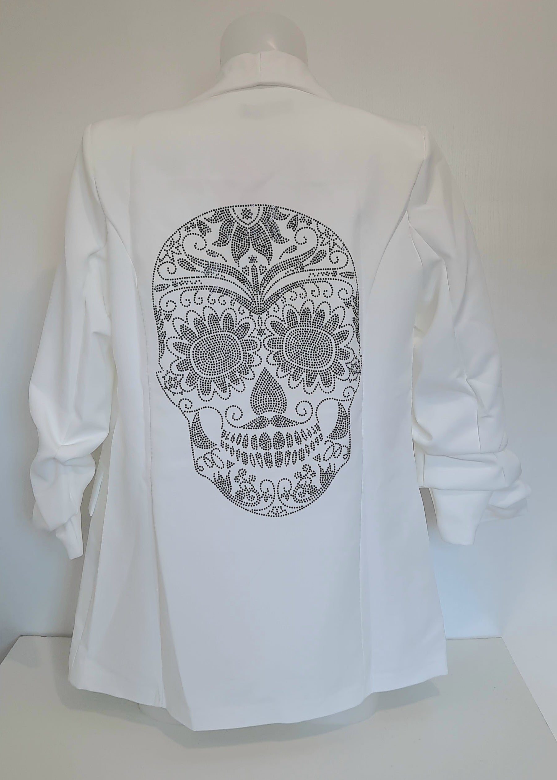 Veste tailleur femme blanche avec coupe droite, manches 3/4 froncées, poches à rabat décoratives et grand motif tête de mort mexicaine en strass noirs dans le dos. Un blazer original et stylé signé Timalya prêt-à-porter féminin.